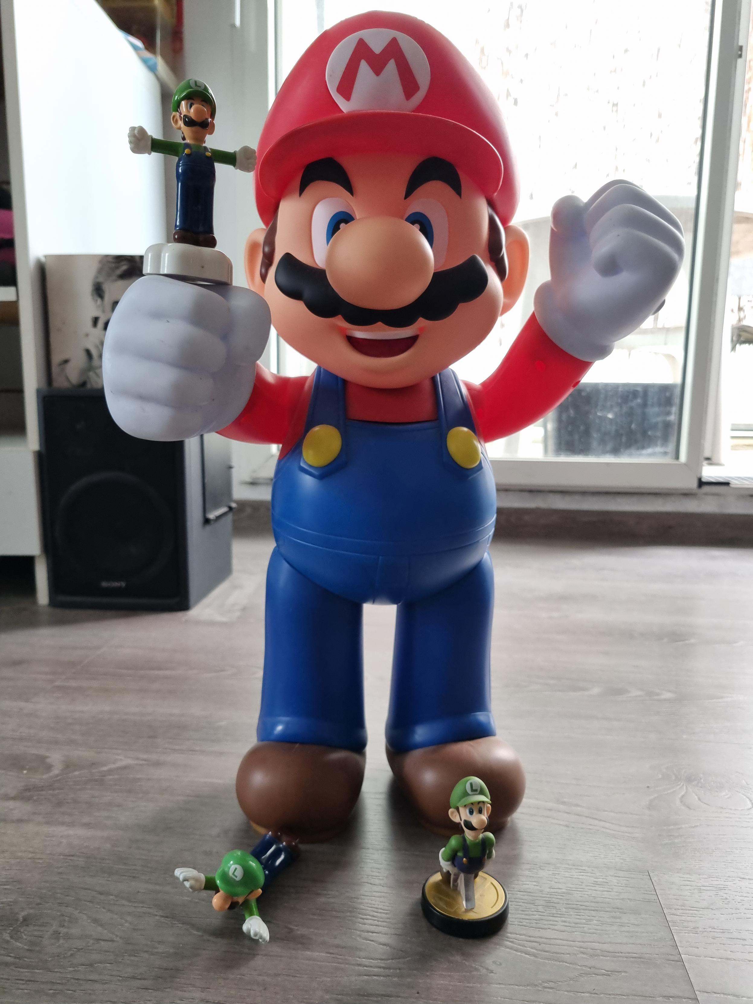 A big posable Mario figurine, with a spinning Luigi resting on a could resting on its first, next to his feet, a figurine of Luigi sliding on the floor, and a Luigi amiibo of him faceplanting